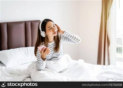 Smile happy young asian woman relaxing and using headphones to listen to music from smart phone in bedroom