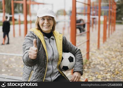 smiley older woman holding football giving thumbs up while working out