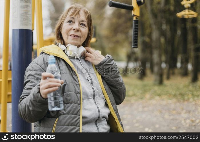 smiley older woman with headphones drinking water after working out outdoors