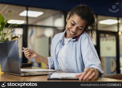 smiley woman office using laptop talking smartphone