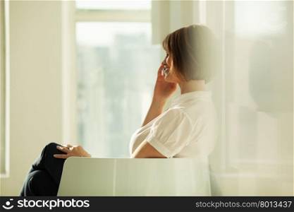 Smiling adult businesswoman talking on smartphone while sitting in chair.. Side view of smiling businesswoman talking on phone