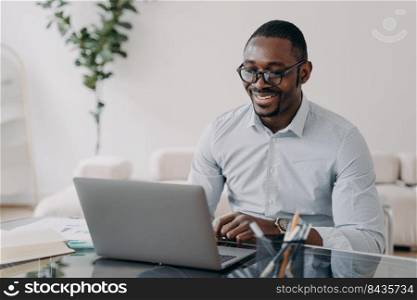 Smiling african american busi≠ssman wearing glasses working at laptop, makes successful deal. Happy black man employee glad to receive good≠ws looking at computer screen at office desk.. Smiling african american man wearing glasses working on busi≠ss project at laptop at office desk