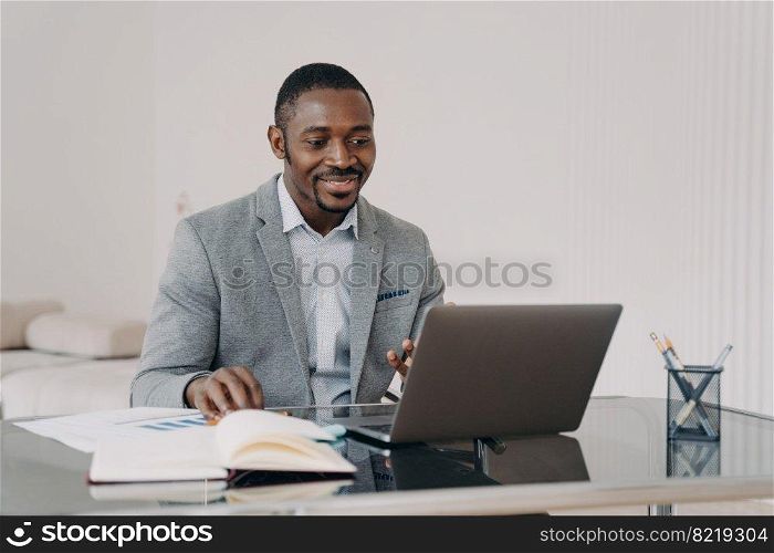 Smiling african american businessman satisfied with his business project, working on laptop, reading email with good news, sitting at office desk. Black male employee watching webinar, learning online. Smiling african american man working on laptop, reading email with good news, sitting at office desk