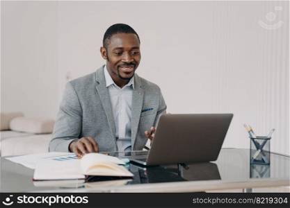 Smiling african american businessman satisfied with his business project, working on laptop, reading email with good news, sitting at office desk. Black male employee watching webinar, learning online. Smiling african american man working on laptop, reading email with good news, sitting at office desk