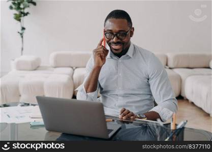 Smiling african american businessman talking on phone looking at laptop at office desk. Black male businessperson marketing and sales manager advising client, makes offer, sells, conducts negotiation. African american businessman talking on phone at laptop advises client, sells, conducts negotiations