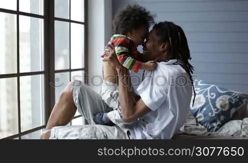 Smiling african american daddy playing with his little toddler boy in the bedroom at home. Joyful african man with dreadlocks holding cute mixed race son and having fun on the bed.
