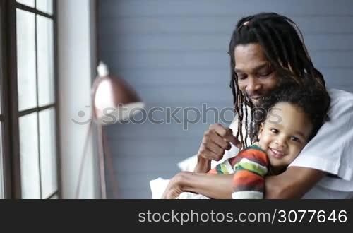 Smiling african american father with dreadlocks embracing his little mixed race toddler son and cheerfully spending time together at home. Handsome man hugging his happy boy with love and tenderness in the bedroom in daylight.