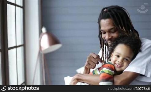 Smiling african american father with dreadlocks embracing his little mixed race toddler son and cheerfully spending time together at home. Handsome man hugging his happy boy with love and tenderness in the bedroom in daylight.