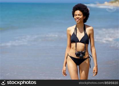 Smiling African American female tourist in bikini with photo camera on neck walking along rippling sea during vacation on sunny summer day. Joyful black woman with photo camera on wet coast