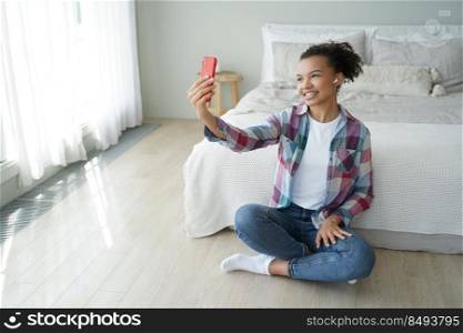 Smiling african american girl raise hand make selfie photo, sitting on the floor in bedroom. Friendly biracial teenager holding smartphone, chatting with friend by video call at home.. African american girl holds phone chatting with friend by video call sitting on the floor in bedroom
