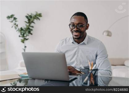 Smiling african american male employee wearing glasses reading email with good news, working on business project at laptop. Modern black businessman manager works, sitting at office desk.. Smiling african american male employee in glasses reading email works on laptop on business project