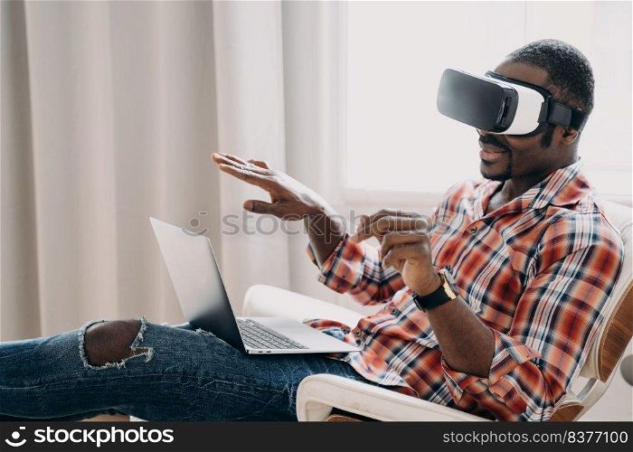 Smiling african american man in VR glasses sits in armchair with laptop, testing virtual reality goggles. Modern black guy viewing purchase in cyberspace. Tech and e-commerce concept.. African american man in virtual reality glasses view purchase online at laptop. Tech, e-commerce