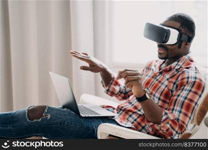 Smiling african american man in VR glasses sits in armchair with laptop, testing virtual reality goggles. Modern black guy viewing purchase in cyberspace. Tech and e-commerce concept.. African american man in virtual reality glasses view purchase online at laptop. Tech, e-commerce