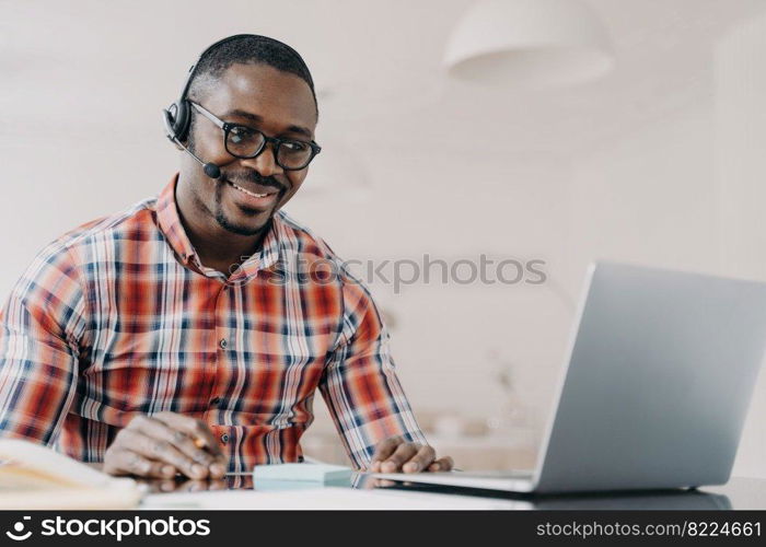 Smiling african american man in wireless headset sits at desk, studying online on laptop, happy modern young black guy in headphones watching webinar on computer. E-learning, distance education.. Smiling african american man in wireless headset sits at desk, studying online on laptop. E-learning