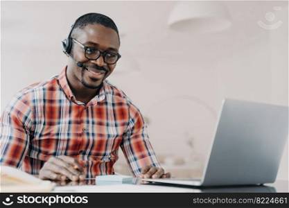 Smiling african american man in wireless headset sits at desk, studying online on laptop, happy modern young black guy in headphones watching webinar on computer. E-learning, distance education.. Smiling african american man in wireless headset sits at desk, studying online on laptop. E-learning