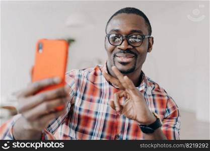 Smiling african american man showing ok approving gesture by hand, holding smartphone, communicates by video call. Modern black guy wearing glasses, looking at phone screen, showing okay sign.. African american man shows ok approving gesture by hand, holding phone, communicates by video call