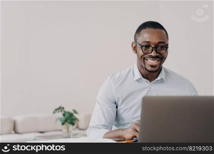 Smiling african american man software developer wearing glasses working on project online at laptop. Happy black guy programmer creating modern application or reading email with good news.. Smiling african american man software developer working online at laptop reads email with good news