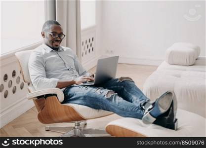 Smiling african american man uses laptop sitting in comfortable chair, black businessman looking at computer screen, communicating on social network, shopping online, working remotely at home.. Smiling african american businessman uses laptop sitting in comfortable chair works remotely at home