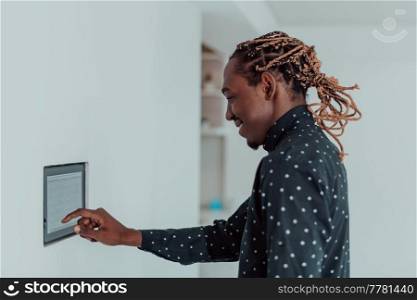 Smiling African American man using modern smart home system, controller on wall, positive young man switching temperature on thermostat or activating security alarm in apartment. High quality photo. Smiling African American man using modern smart home system, controller on wall, positive young man switching temperature on thermostat or activating security alarm in apartment