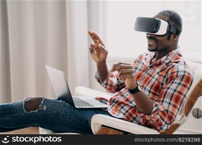 Smiling african american man wearing virtual reality glasses working in cyberspace or playing video game on laptop, sitting in chair. modern black male software developer testing VR application.. African american man in virtual reality glasses works or play video game on laptop, sitting in chair