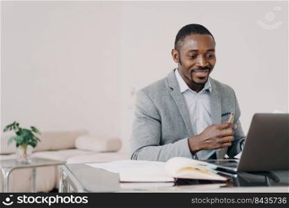 Smiling african american man working at laptop online at office desk. Black male businessperson looking at computer screen, reading email with good news about profitable deal or job promotion.. African american businessman reading email with good news at laptop at office desk, smiling