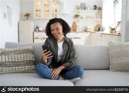 Smiling african american teen girl chatting in social network, using modern mobile phone apps, sitting on cozy couch at home. Happy mixed race young lady holding smartphone, shopping in online store.. African american teen girl use modern phone apps chatting in social network, shopping online at home