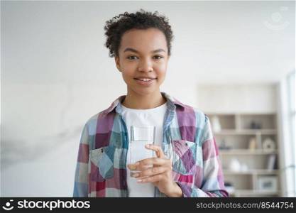 Smiling african american teen girl holds glass with mineral clean water standing at home. Happy young woman looking at camera holding fresh pure aqua. Healthy lifestyle habit, wellness concept.. African american girl holds glass of purified water standing at home. Healthy lifestyle, wellness