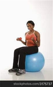 Smiling African American young adult woman sitting on exercise ball holding dumbbells.