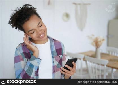 Smiling african american young female holding smartphone, touching wireless earphones in ear, listening to music, songs on radio at home. Happy biracial teen girl enjoying using musical apps on phone.. African american young girl listens to music using smartphone at home. Musical apps advertising