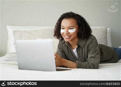 Smiling african american young girl with under eye patches on face works at laptop, lying on bed. Biracial woman with moisturizing cotton pads watching series. Morning facial skincare routine, beauty.. Biracial girl with under eye patches on face work at laptop. Morning facial skincare routine, beauty