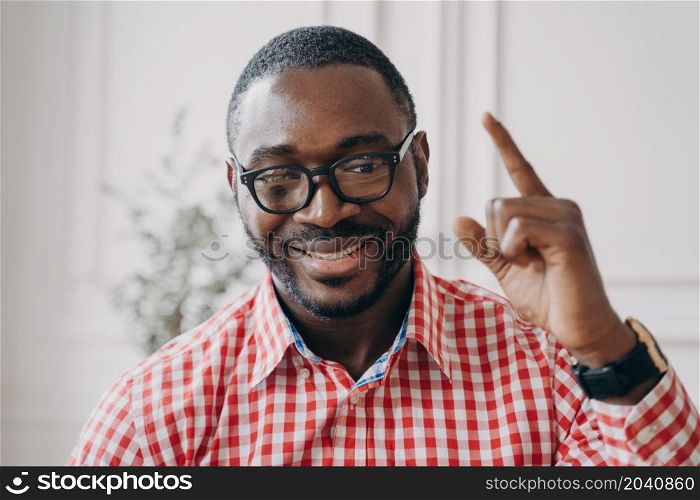 Smiling African ethnicity male English tutor in glasses raises hand points out with index finger up while explaining homework to students online. Cheerful businessman talking with collegues during videocall. Smiling African ethnicity male English tutor in glasses raises hand points out with index finger up