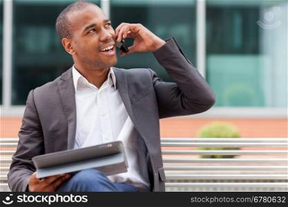 Smiling afro american manager sitting on a bench and phoning