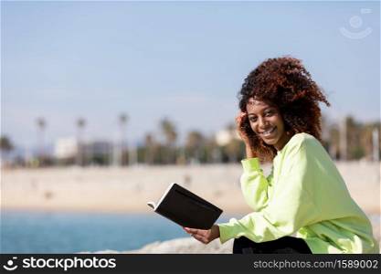 Smiling afro american woman sitting on shore while reading a book in a sunny day
