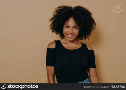 Smiling afro american woman with curly hair standing against beige background, has happy and positive face expression, young african female in casual outfit looking at camera with tender smile. Smiling afro american woman with curly hair standing against beige background