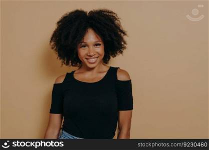 Smiling afro american woman with curly hair standing against beige background, has happy and positive face expression, young african female in casual outfit looking at camera with tender smile. Smiling afro american woman with curly hair standing against beige background
