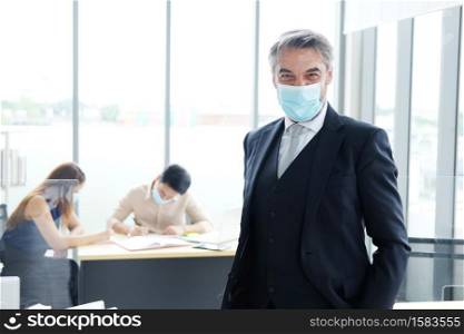 Smiling and Smart Caucasian senior businessman in black suit and asian staff and teamwork wearing masks prevent covid 19 virus at office. New nomal and social distace of business concept.