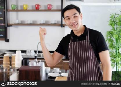Smiling Asian barista young man is Thumbs up for feeling proud of the coffee shop is start up business. Successful businessman is owner of cafe.