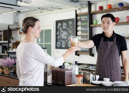 Smiling Asian barista young man serving a glass of iced latte coffee for Caucasian woman customer at counter bar in coffee shop. 