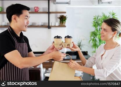 Smiling Asian barista young man serving two glass of hot coffee and holding paper bags and take away to home for Caucasian woman customer at counter bar in coffee shop.