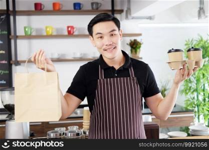 Smiling Asian barista young man serving two glass of hot coffee and holding paper bags and take away to home for customers at counter bar in coffee shop. Start up for bussiness Concept