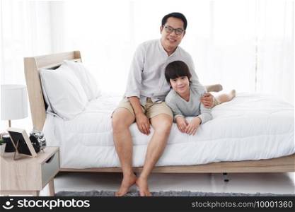 Smiling Asian cute little boy is relaxing and hugging with father in bedroom at home. Family love and child educational for homeschool on holiday concept.