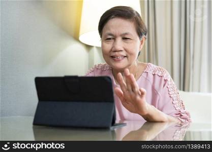 Smiling Asian Elderly woman making video call on digital tablet during quarantine and isolated at home. Happy Senior female in pink cloth waving hand while talking with people via internet technology.