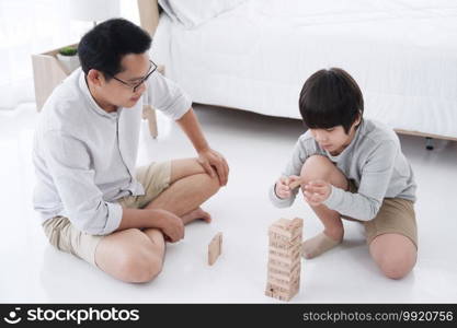 Smiling Asian father and cute little boy is enjoying and funny with wooden blocks game together in bedroom on holiday. Family activities and child educational for homeschool concept.