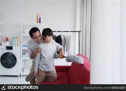 Smiling Asian father and little boy child is enjoying for House cleaning together in Laundry room. They is Dust off the red sofa  by Feather duster. Homeschool and Family time on holiday concept