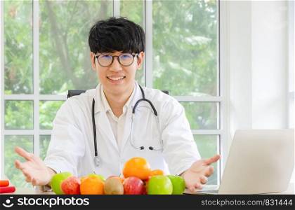 Smiling asian male nutritionist wearing glasses in lab coat uniform looking at camera and holding healthy fresh fruits of apple, orange and kiwifruit on the desk in his office. Hhealthcare and diet concept.
