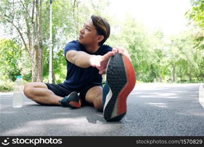 Smiling Asian man sitting on concrete floor for exercising and stretching the legs to relax the muscles after running and jogging in the garden. Sport and healthy Concept