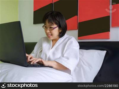 Smiling Asian middle aged woman in white bathrobe using laptop computer on pillow in her bed at morning time, cozy relaxation concept
