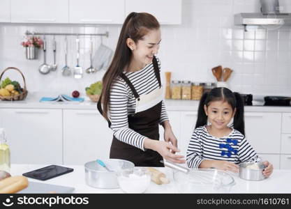 Smiling Asian mother and little asian girl child is cooking for baking bakery on wooden table together in kitchen. Homemade pastry and mix ingredient for bread. Family love and Homeschool Concept.