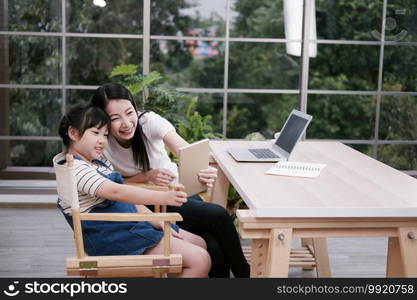 Smiling Asian mother and little girl child is selfie by tablet and online learning with laptop together in living room. Homeschool and educational with technology concept