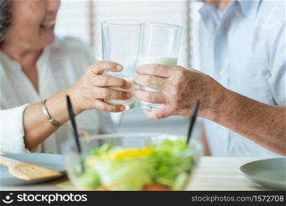 Smiling Asian senior man and woman holding glasses of milk. Cheerful Elderly couple enjoying healthy salad food while having breakfast at home. Happy Lifestyle Grandfather and Grandmother.